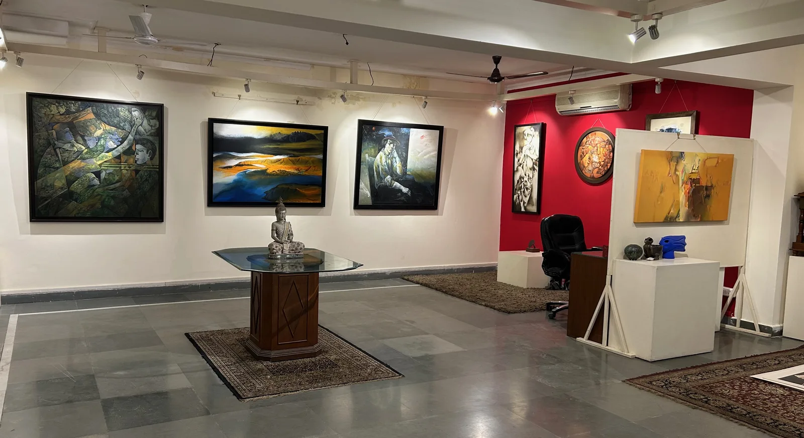 Gallery One — main exhibition hall with paintings on white walls, red accent wall, central display table with Buddha sculpture, and sculpture plinths